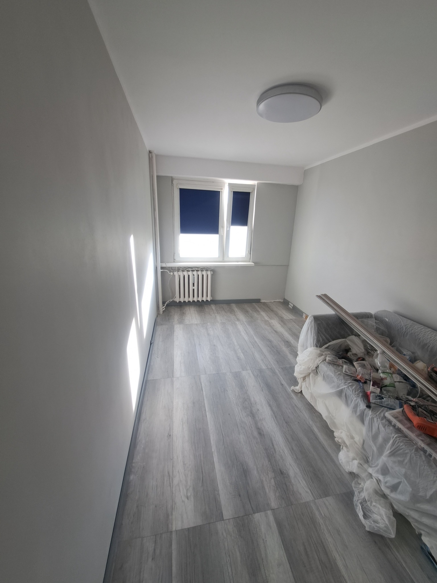 Pokój po świeżym malowaniu na szaro, z nową podłogą imitującą drewno, okno z zasłoniętą roletą i zakryty folią mebel z narzędziami malarskimi.