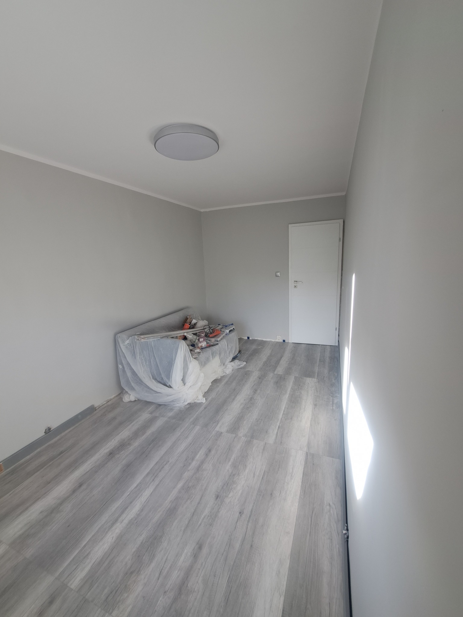 Pokój po odświeżeniu, ściany pomalowane na szaro, podłoga wyłożona panelami imitującymi drewno, zabezpieczona folią kanapa z narzędziami malarskimi.