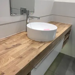 Nowoczesna łazienka z okrągłą, białą umywalką nablatową na blacie z drewna dębowego, chromowana bateria, białe szafki pod umywalką i szara podłoga.