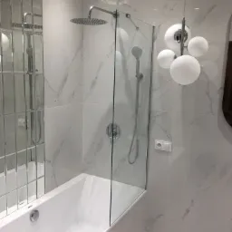 Biała wanna z szklanym parawanem nawannowym, deszczownicą i baterią podtynkową na tle marmurowych płytek, oświetlona nowoczesną lampą kulistą.