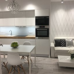 KuchnieNaWymiar - Biała kuchnia z drewnianym blatem, widok na stół z owocami i salon z sofą na tle dekoracyjnej ściany.