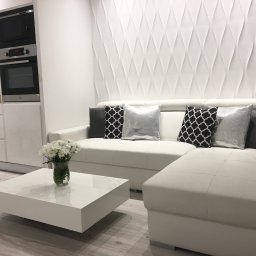 KuchnieNaWymiar - Jasne wnętrze: biała sofa narożna z dekoracyjnymi poduszkami, minimalistyczny stolik kawowy z bukietem białych kwiatów, zabudowa kuchenna z piekarnikiem i mikrofalówką oraz panel ścienny 3D.