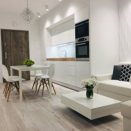 KuchnieNaWymiar - Jasne wnętrze: biała kuchnia z zabudową, stół z owocami, sofa z poduszkami, dekoracyjna ściana i drewnopodobna podłoga.