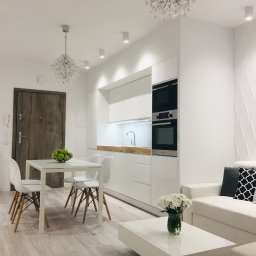 KuchnieNaWymiar - Jasne wnętrze mieszkania z białą kuchnią, stołem z jabłkami, sofą i kryształowymi żyrandolami, drewniana podłoga i drzwi.