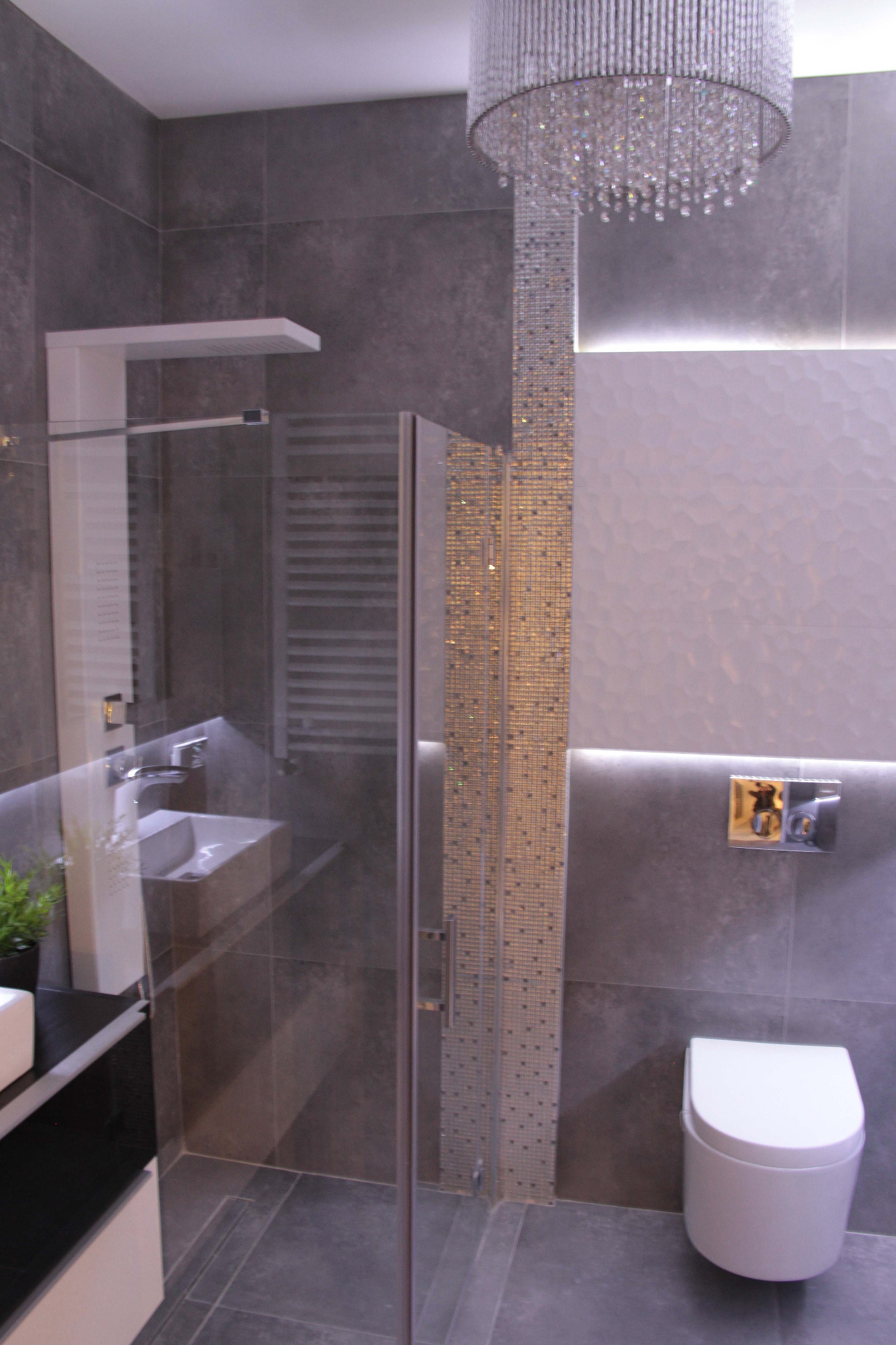 Nowoczesna łazienka z szarą mozaiką, kabiną prysznicową ze szklanymi drzwiami, wiszącą toaletą i designerskim żyrandolem.