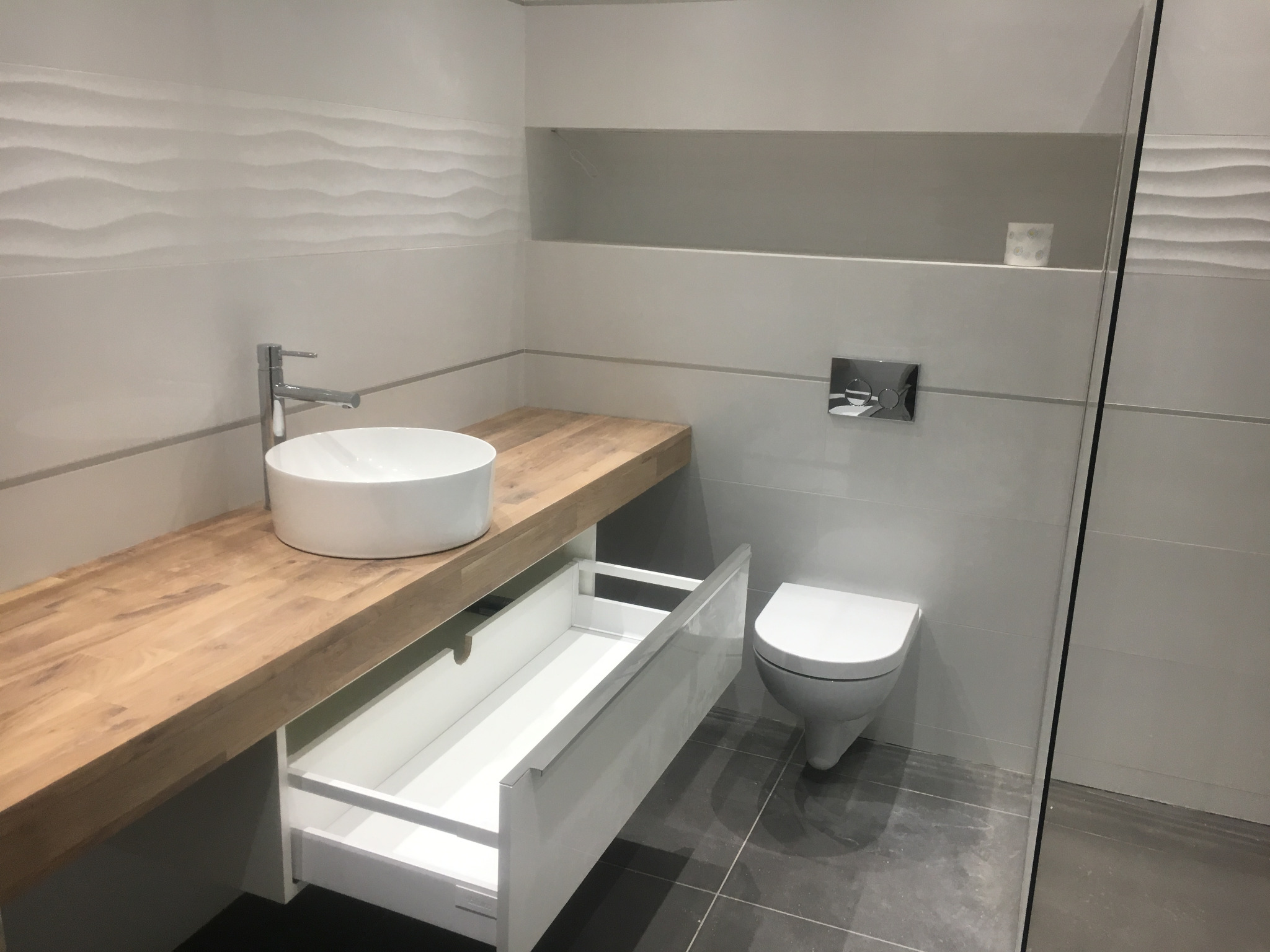 Nowoczesna łazienka z drewnianym blatem, okrągłą umywalką nablatową i otwartą szufladą pod blatem, minimalistyczna toaleta podwieszana.
