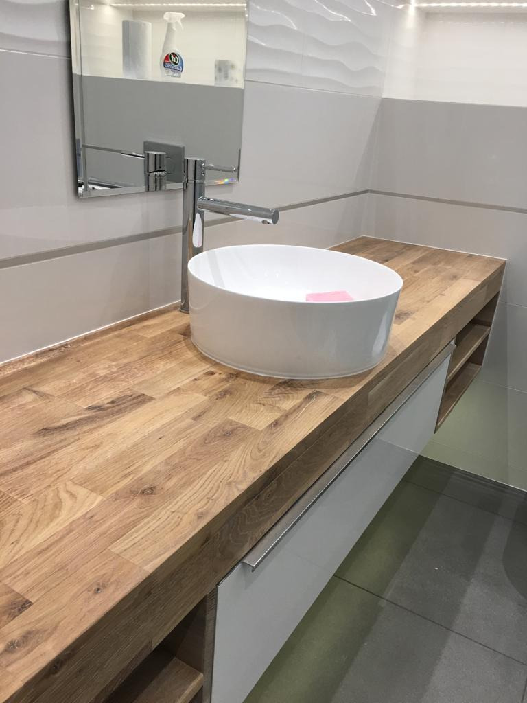 Nowoczesna łazienka z okrągłą, białą umywalką nablatową na blacie z drewna dębowego, chromowana bateria, białe szafki pod umywalką i szara podłoga.