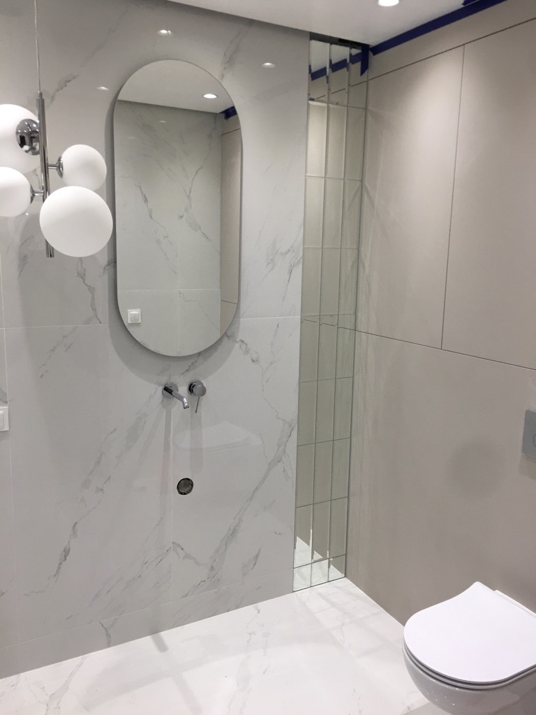 Nowoczesna łazienka z marmurowymi płytkami, owalnym lustrem nad umywalką i pionowymi lustrzanymi panelami, podkreślona designerską lampą kulistą.