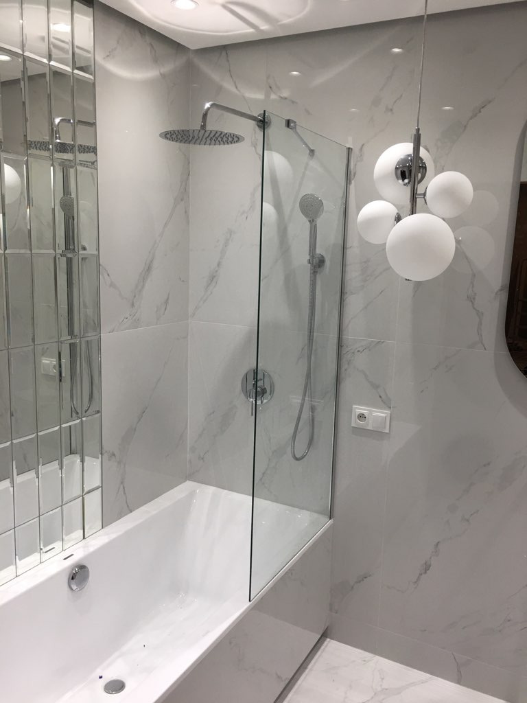 Biała wanna z szklanym parawanem nawannowym, deszczownicą i baterią podtynkową na tle marmurowych płytek, oświetlona nowoczesną lampą kulistą.