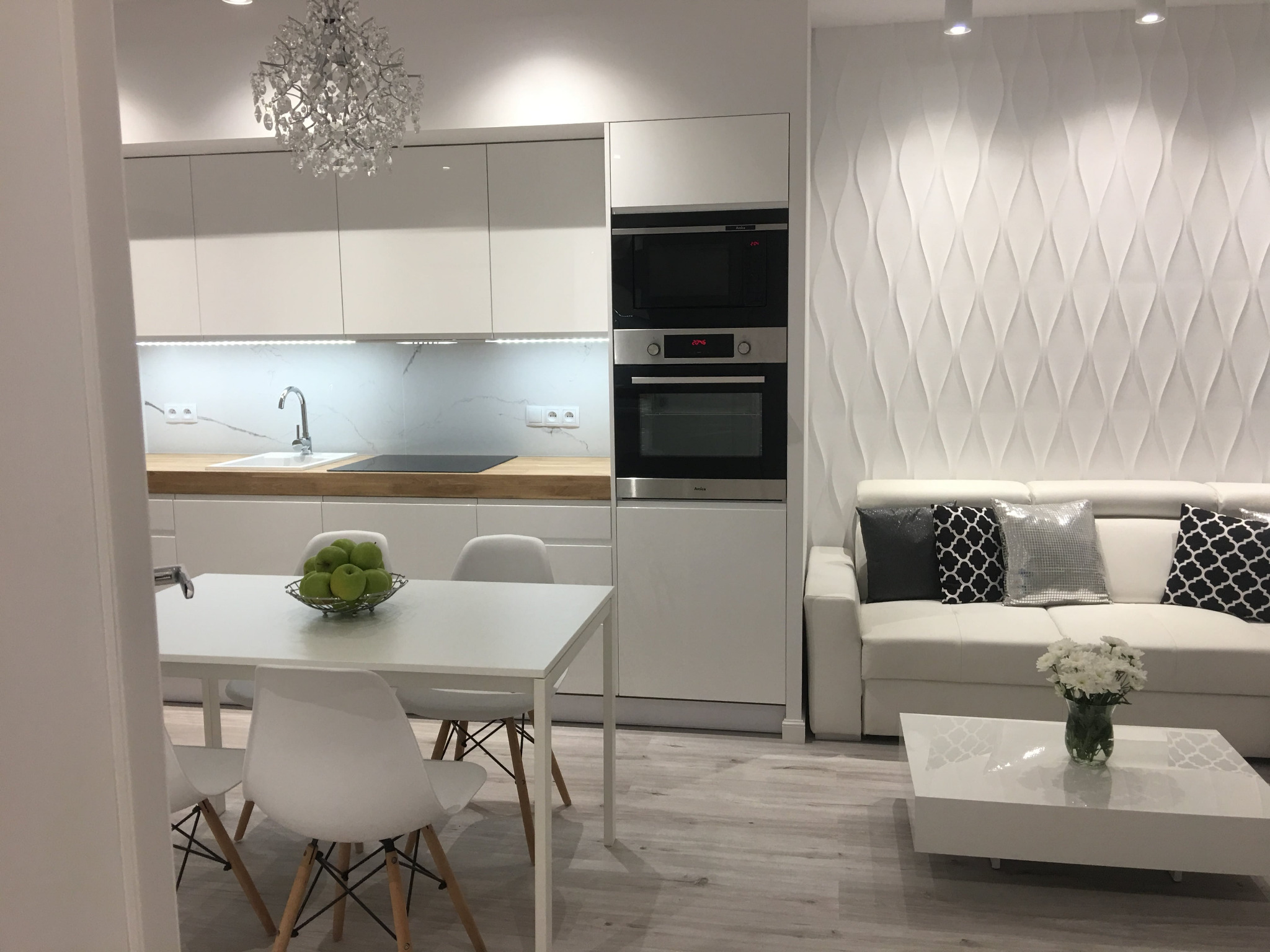 Biała kuchnia z drewnianym blatem, widok na stół z owocami i salon z sofą na tle dekoracyjnej ściany.