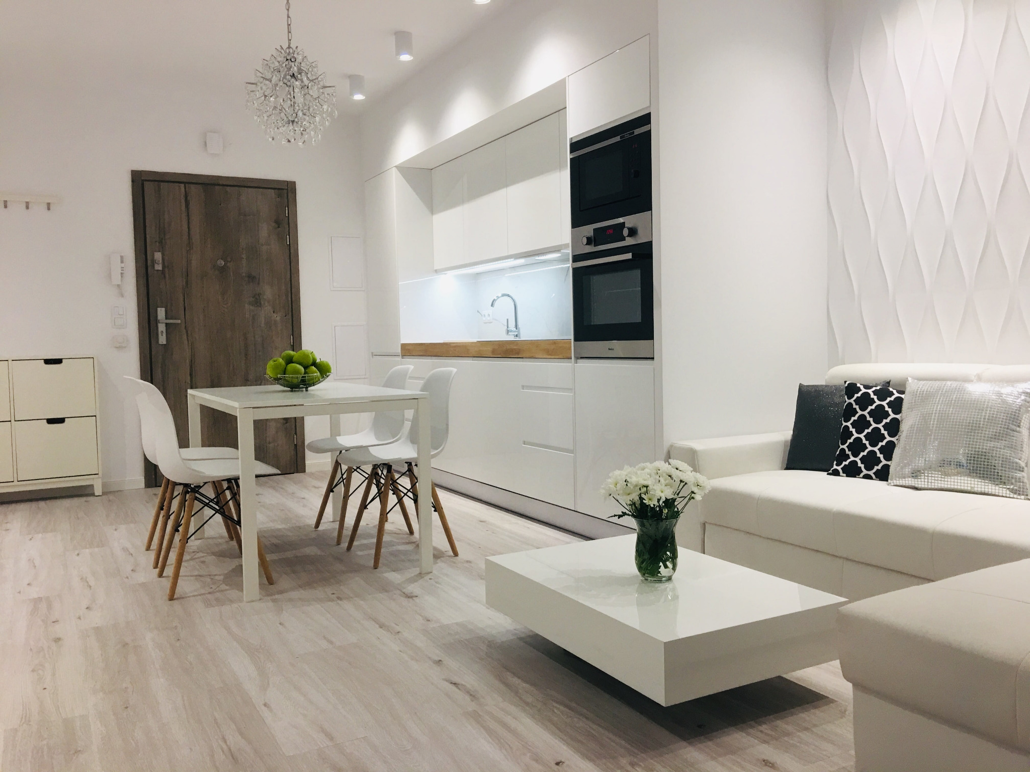 Jasne wnętrze: biała kuchnia z zabudową, stół z owocami, sofa z poduszkami, dekoracyjna ściana i drewnopodobna podłoga.