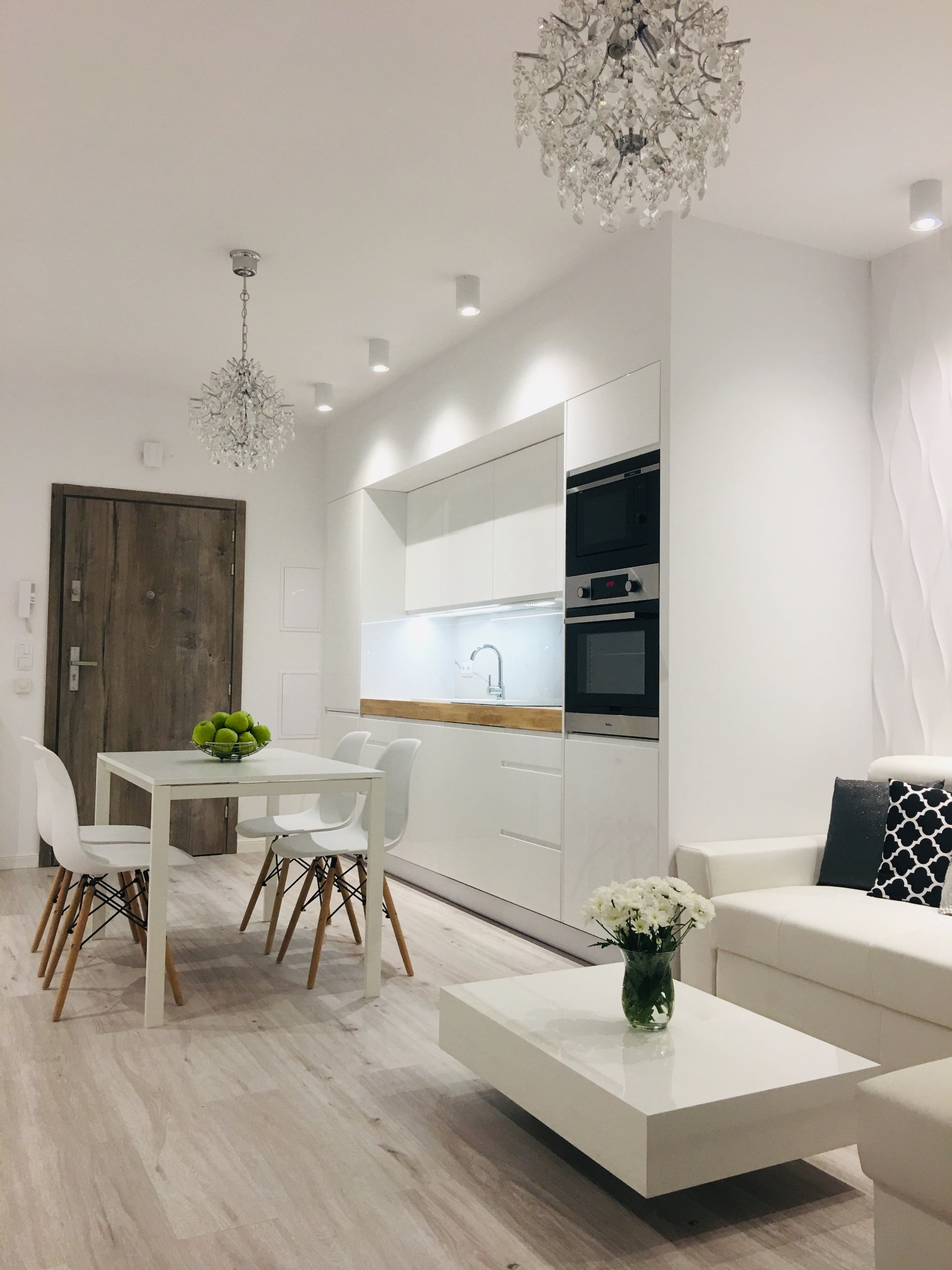 Jasne wnętrze mieszkania z białą kuchnią, stołem z jabłkami, sofą i kryształowymi żyrandolami, drewniana podłoga i drzwi.
