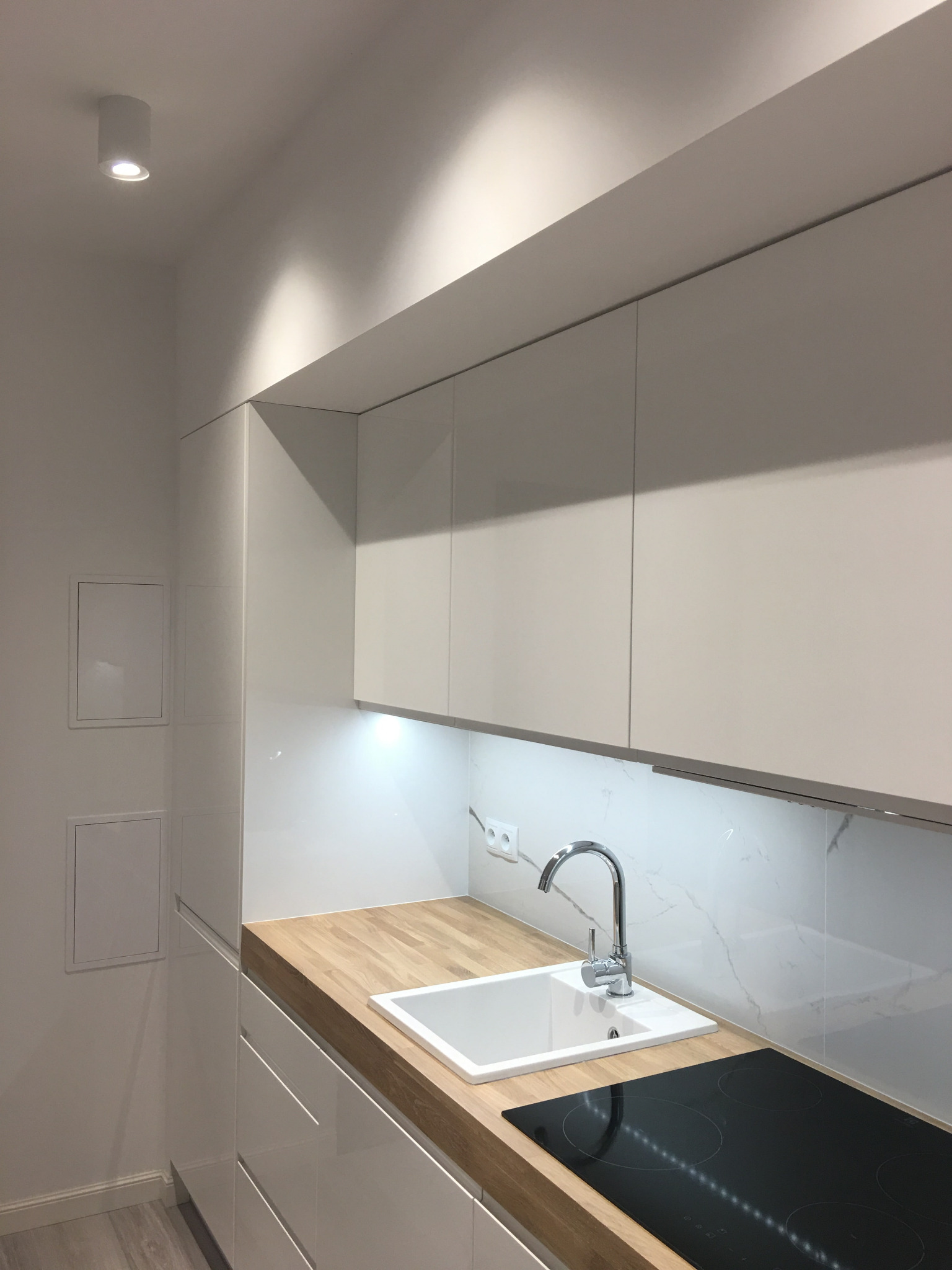 Nowoczesna, minimalistyczna kuchnia: białe, lakierowane fronty szafek, drewniany blat, biały zlew podwieszany i indukcyjna płyta grzewcza.