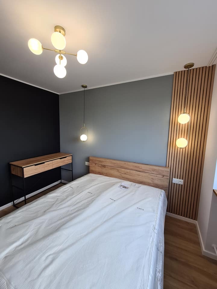 Nowoczesna sypialnia z łóżkiem z drewnianym zagłówkiem, biurkiem na metalowych nogach, szarą ścianą, czarną ścianą akcentującą oraz oświetleniem: lampą sufitową z kulistymi kloszami i pionowym...