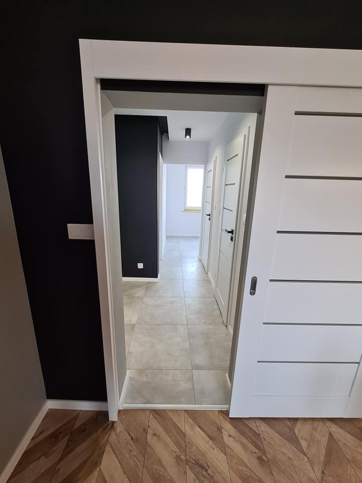 Zamknięte białe drzwi przesuwne z poziomymi frezami, widok na korytarz z szarą podłogą z płytek i białymi drzwiami, czarna ściana po lewej stronie, parkiet ułożony w jodełkę na podłodze.