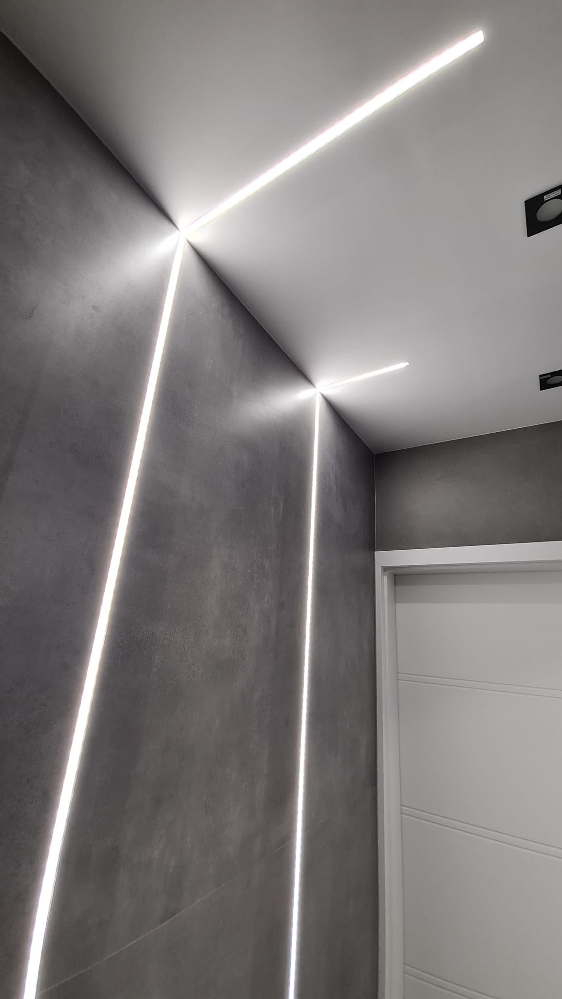 Szara ściana z dekoracyjnym oświetleniem LED w formie pionowych i poziomych linii, białe drzwi w tle.