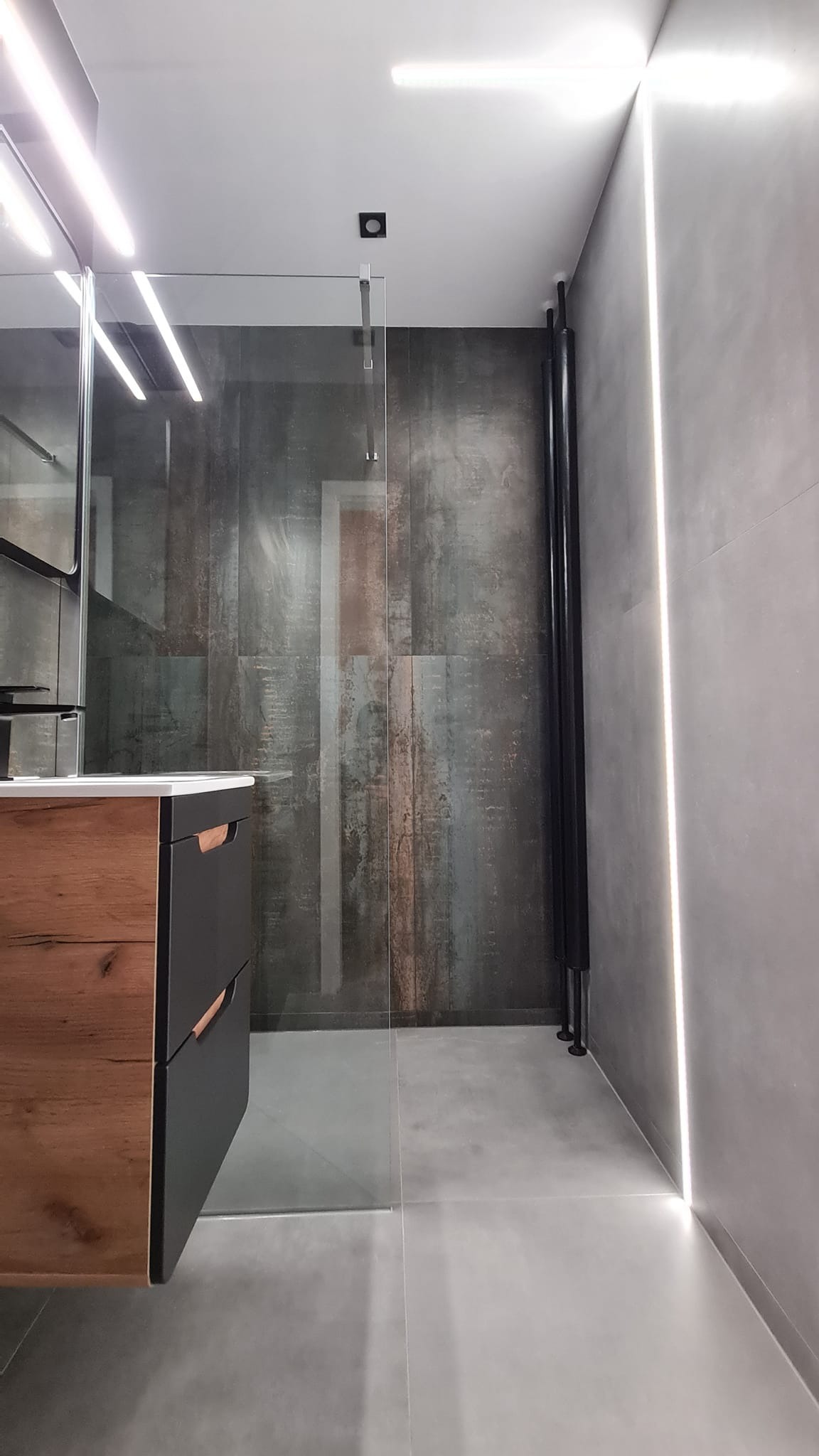 Nowoczesna łazienka z kabiną prysznicową typu walk-in, drewnianą szafką pod umywalką i pionowym grzejnikiem drabinkowym w kolorze czarnym, oświetlona listwami LED.