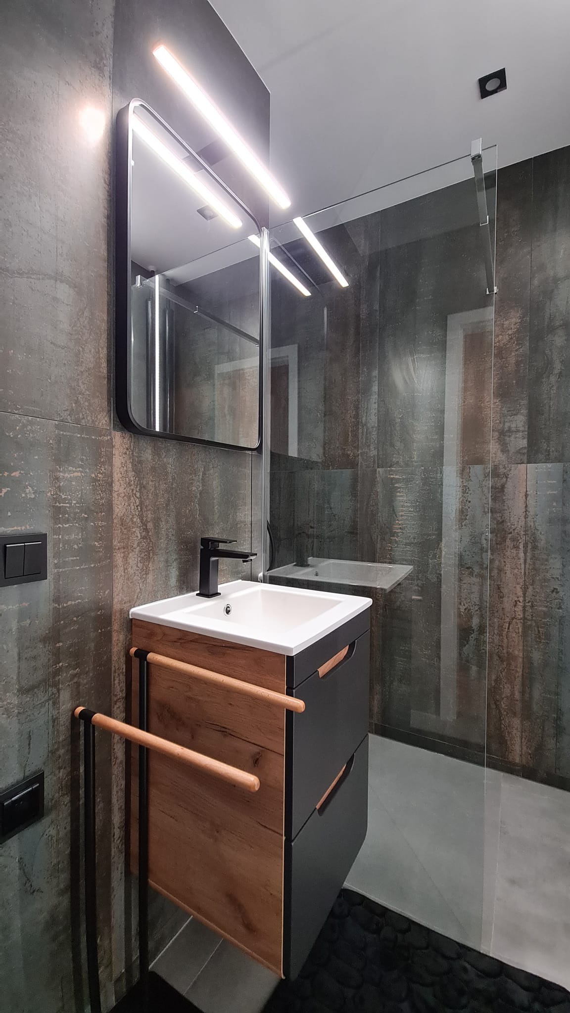 Nowoczesna łazienka z umywalką nablatową, czarną armaturą, szafką z drewnianymi frontami i szklaną kabiną prysznicową.