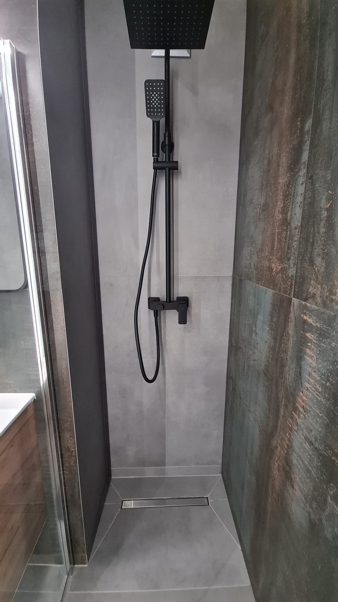 Nowoczesny prysznic typu walk-in z czarną armaturą i odpływem liniowym, wykończony szarą płytką imitującą beton i dekoracyjną płytką z efektem rdzy.
