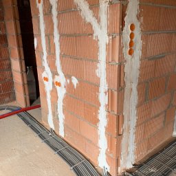 KliMal instalacje - Murowana ściana z widocznymi bruzdami wypełnionymi gipsem, pomarańczowe puszki elektryczne i kable w szarych peszlach przymocowane do podłoża metalowymi uchwytami.