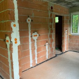 KliMal instalacje - Surowy mur z czerwonej cegły z przygotowanymi otworami pod instalację elektryczną, zabezpieczonymi pomarańczowymi puszkami, oraz rozprowadzonymi przewodami w czarnych peszlach wzdłuż podłogi.