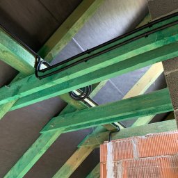 KliMal instalacje - Instalacja elektryczna w peszlu, poprowadzona wzdłuż zielonych belek więźby dachowej, widok z dołu na poddaszu w trakcie budowy.