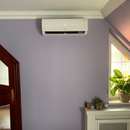 KliMal instalacje - Biały klimatyzator ścienny z wyświetlaczem temperatury, zamontowany na fioletowej ścianie w pokoju z drewnianymi drzwiami i oknem ze szprosami, obok grzejnika dekoracyjnego z roślinami i ozdobami.