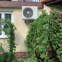 KliMal instalacje - Biały klimatyzator Rotenso zamontowany na beżowej ścianie domu, otoczony bujną zielenią i kwitnącymi różowymi kwiatami, z balkonem z kutą balustradą widocznym powyżej.