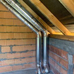 KliMal instalacje - Dwa pionowe, metalowe przewody wentylacyjne, częściowo owinięte folią izolacyjną, w rogu pomieszczenia z surową cegłą i drewnianą konstrukcją dachu.
