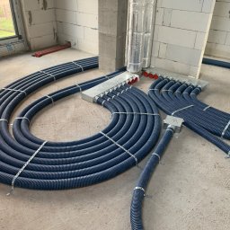 KliMal instalacje - Niebieskie, karbowane rury instalacyjne ułożone w pętle na surowym betonowym podłożu wewnątrz budynku w trakcie budowy, przymocowane metalowymi opaskami, z widocznymi puszkami rozdzielczymi...