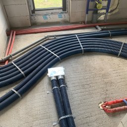 KliMal instalacje - Wewnętrzne instalacje w surowym stanie: wiązka niebieskich rur karbowanych przymocowana metalowymi obejmami do ściany z betonu komórkowego, obok czerwone rury ogrzewania podłogowego i okno...