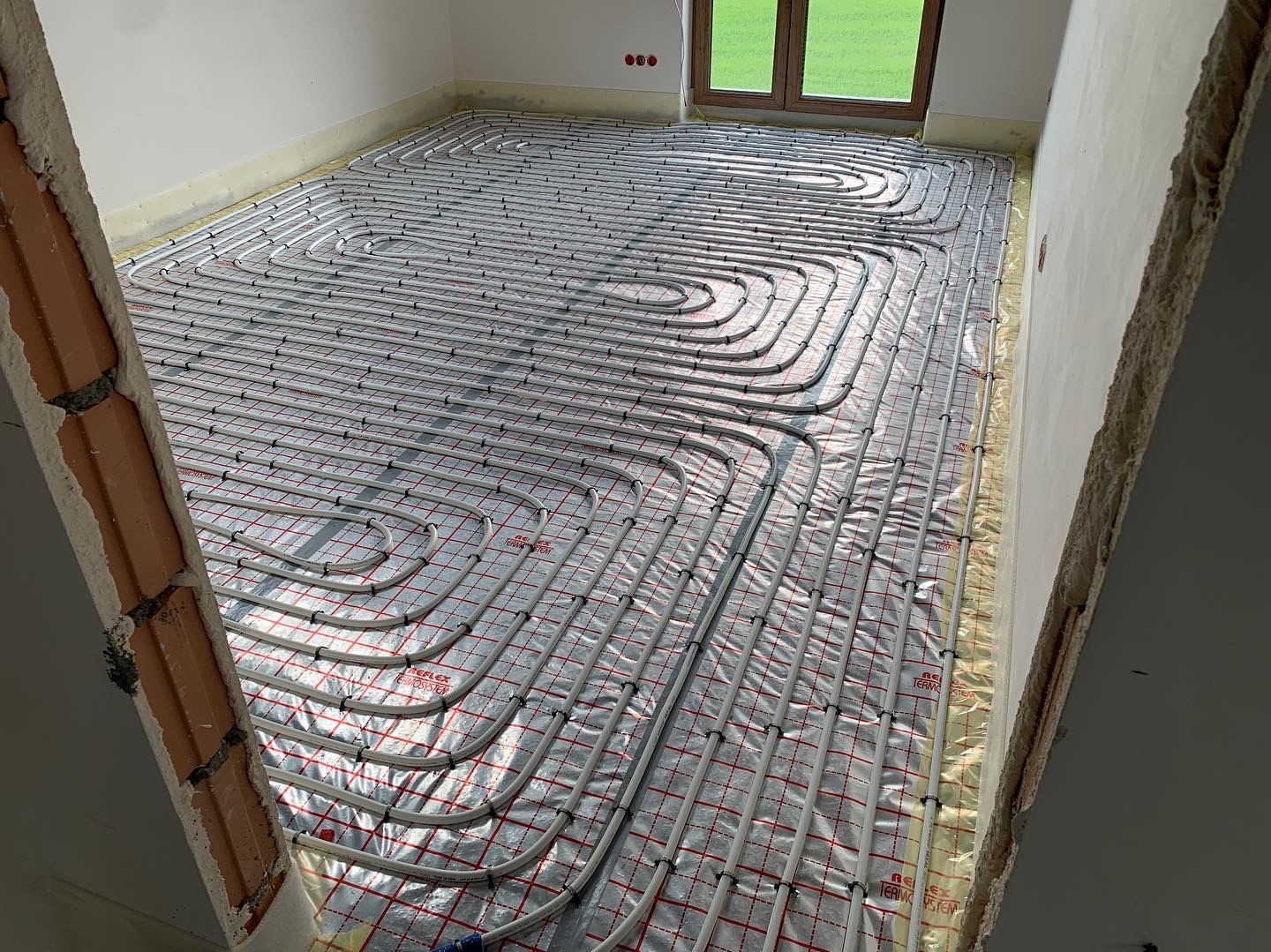 Rozłożona instalacja ogrzewania podłogowego na folii termoizolacyjnej w surowym pomieszczeniu, widoczne rury w regularnych odstępach i fragment ściany w stanie deweloperskim.