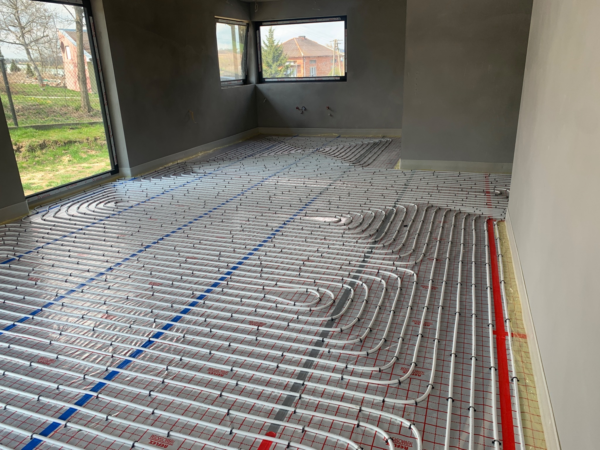 Rozłożona instalacja ogrzewania podłogowego w surowym wnętrzu z widokiem na zewnątrz przez okna.