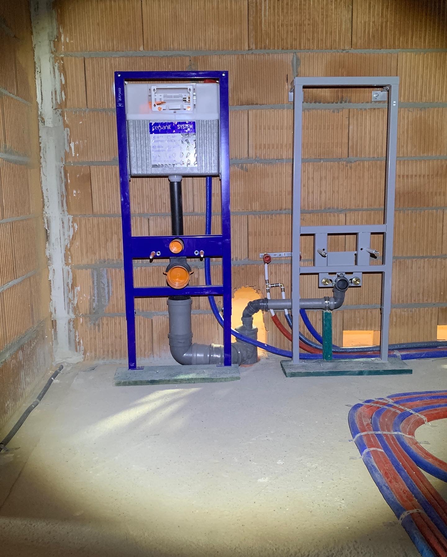 Dwa stelaże podtynkowe do WC, jeden w kolorze niebieskim z widoczną spłuczką Cersanit System, drugi szary, oba zamontowane na surowej ścianie z cegły, z widocznymi rurami doprowadzającymi wodę...