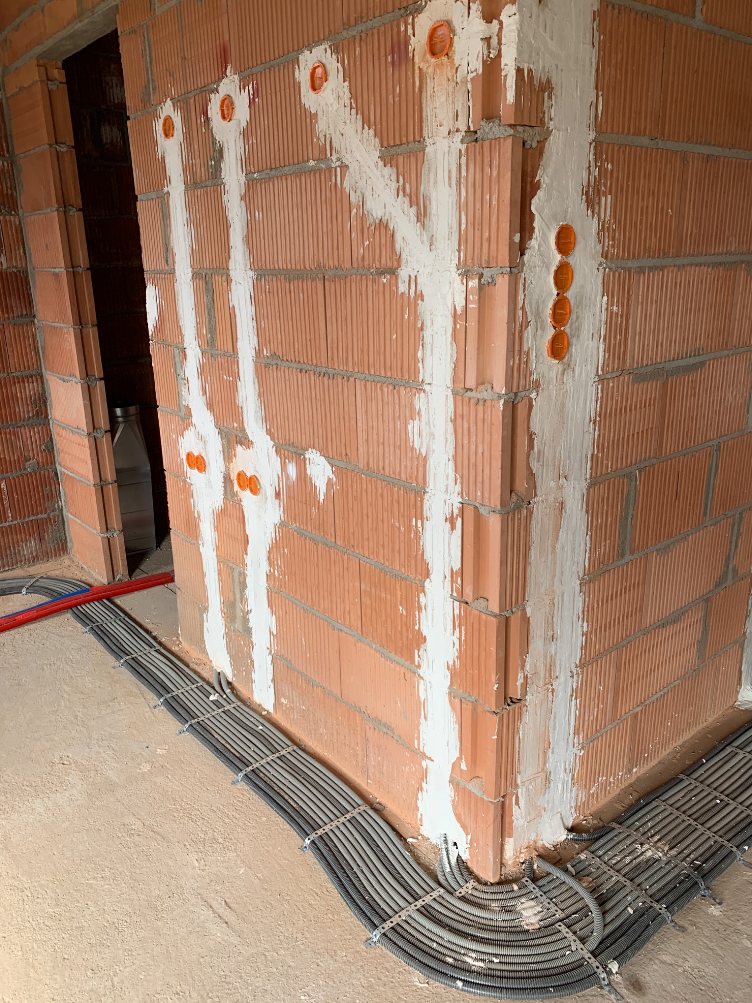 Murowana ściana z widocznymi bruzdami wypełnionymi gipsem, pomarańczowe puszki elektryczne i kable w szarych peszlach przymocowane do podłoża metalowymi uchwytami.