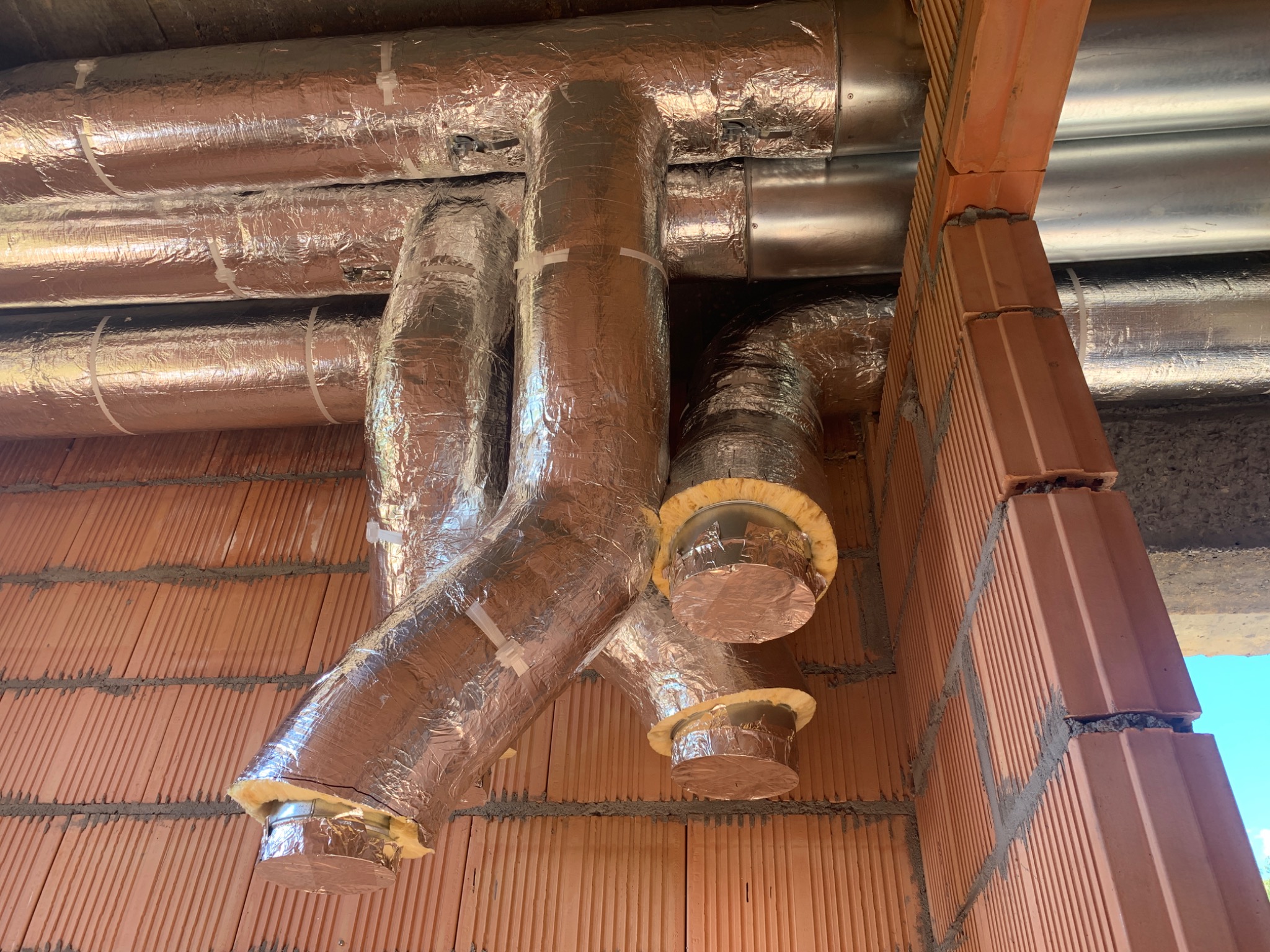 System kanałów wentylacyjnych owiniętych folią aluminiową, zamontowanych pod sufitem z cegieł, widoczne izolacje z wełny mineralnej na końcach kanałów.