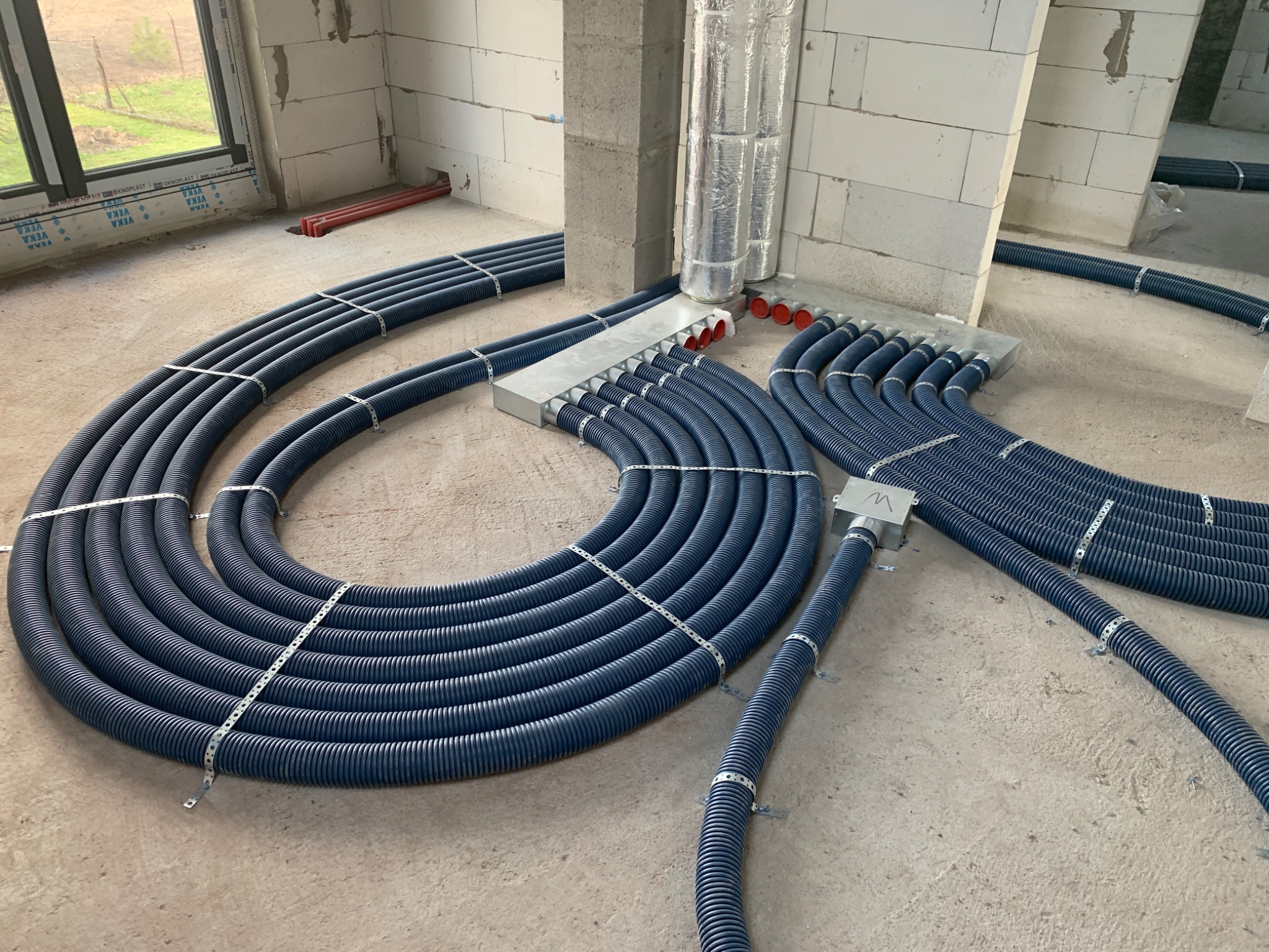 Niebieskie, karbowane rury instalacyjne ułożone w pętle na surowym betonowym podłożu wewnątrz budynku w trakcie budowy, przymocowane metalowymi opaskami, z widocznymi puszkami rozdzielczymi...