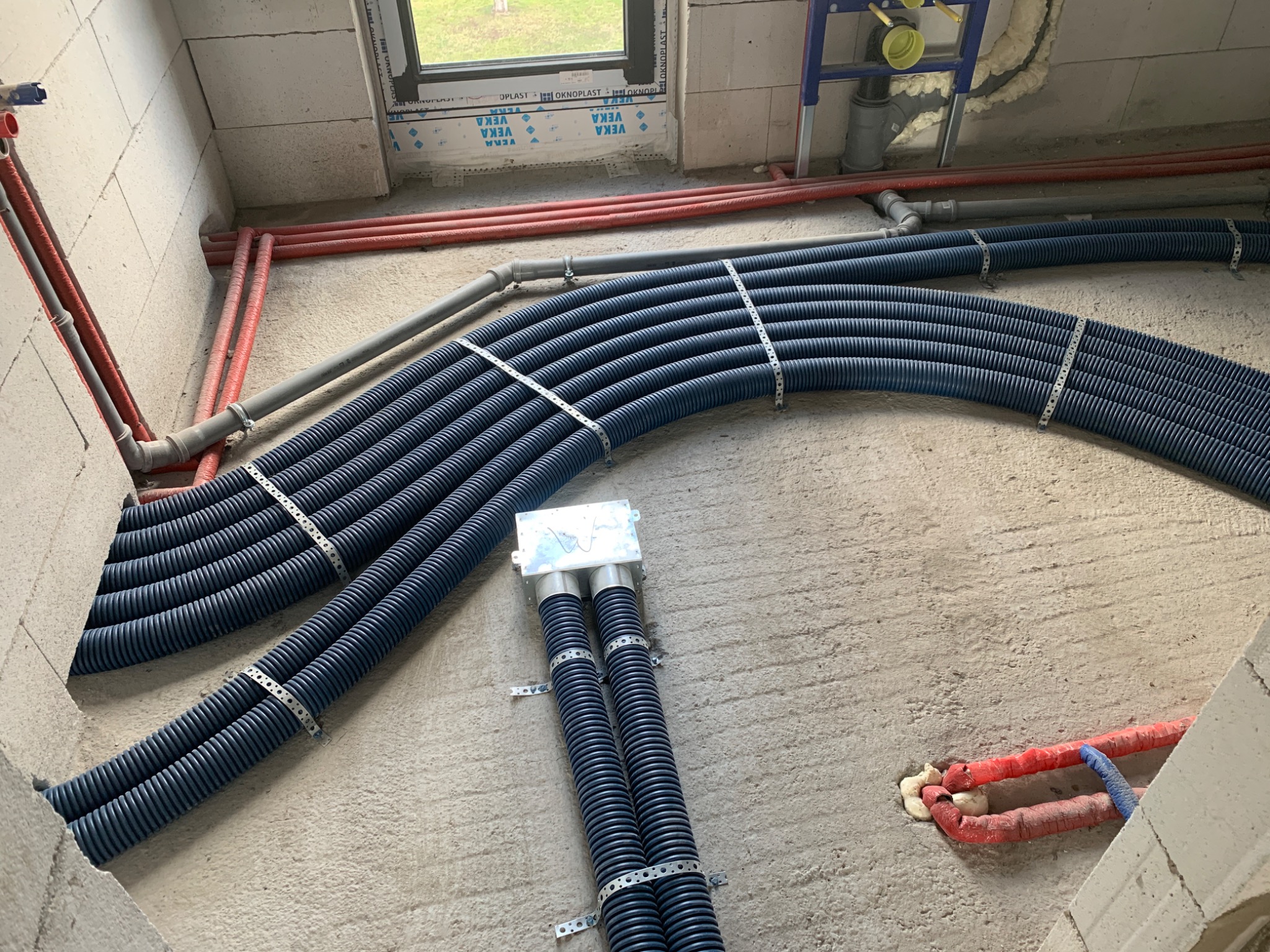 Wewnętrzne instalacje w surowym stanie: wiązka niebieskich rur karbowanych przymocowana metalowymi obejmami do ściany z betonu komórkowego, obok czerwone rury ogrzewania podłogowego i okno...