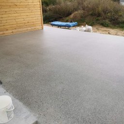 Krzysztof Rog - Świeżo wylana, szlifowana betonowa powierzchnia na zewnątrz budynku z drewnianą elewacją. W tle widoczne worki z materiałami budowlanymi i przykryte meble ogrodowe.