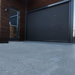 Krzysztof Rog - Szara, gładka powierzchnia betonowa przed budynkiem z drewnianą elewacją i balkonem z metalową balustradą.