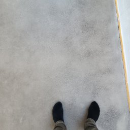Krzysztof Rog - Szara, gładka powierzchnia podłogi z widocznymi stopami w czarnych skarpetach i spodniami khaki. Wzdłuż krawędzi widoczna żółta taśma malarska zabezpieczająca ścianę.