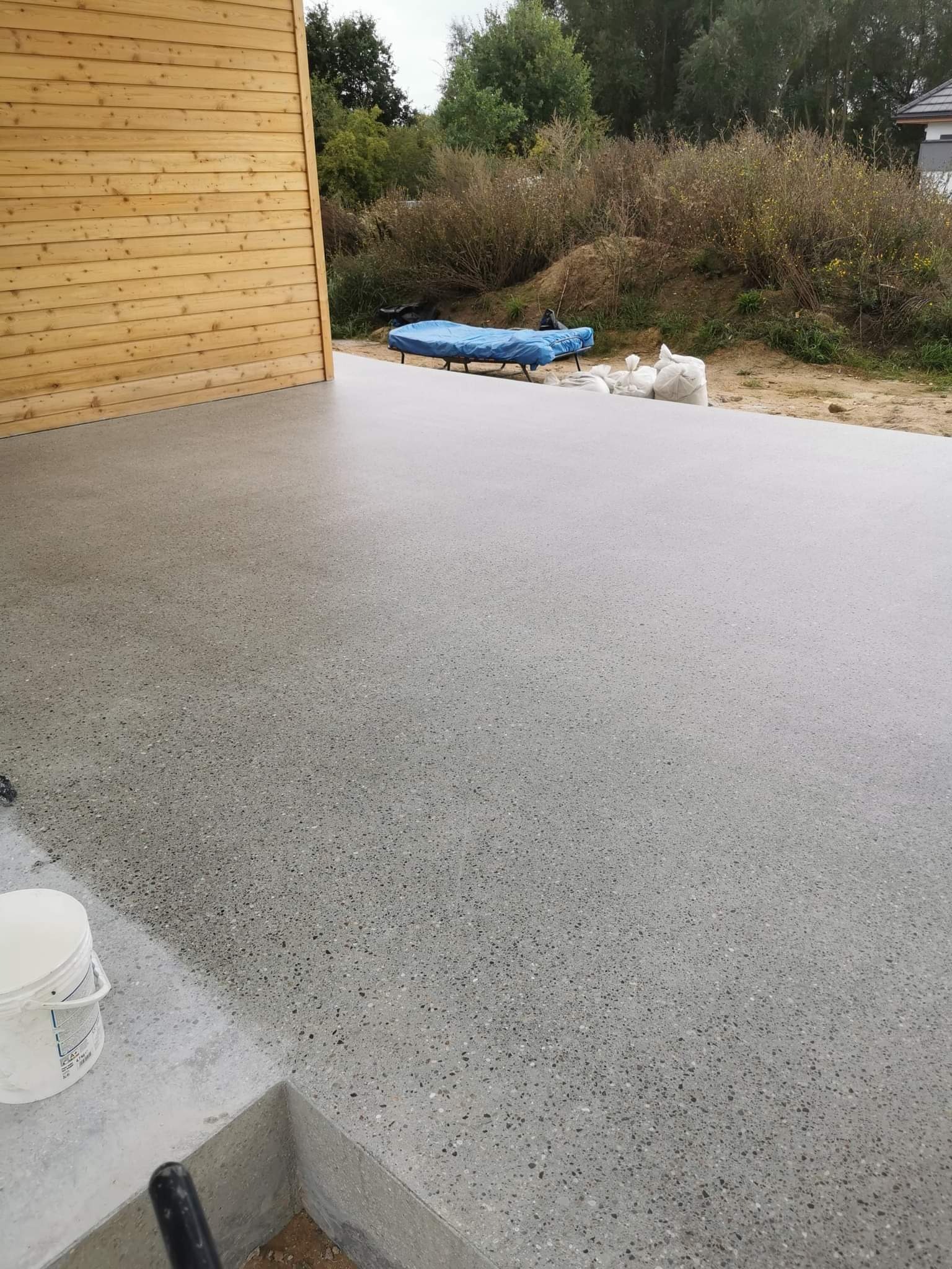 Świeżo wylana, szlifowana betonowa powierzchnia na zewnątrz budynku z drewnianą elewacją. W tle widoczne worki z materiałami budowlanymi i przykryte meble ogrodowe.