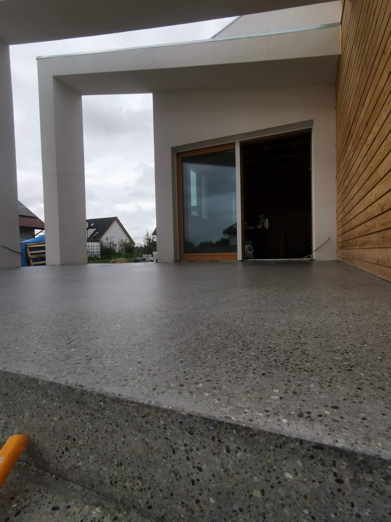 Szlifowana, betonowa powierzchnia tarasu z widocznymi ziarnami kruszywa, nowoczesny budynek w tle z drewnianą elewacją i przesuwnymi drzwiami.