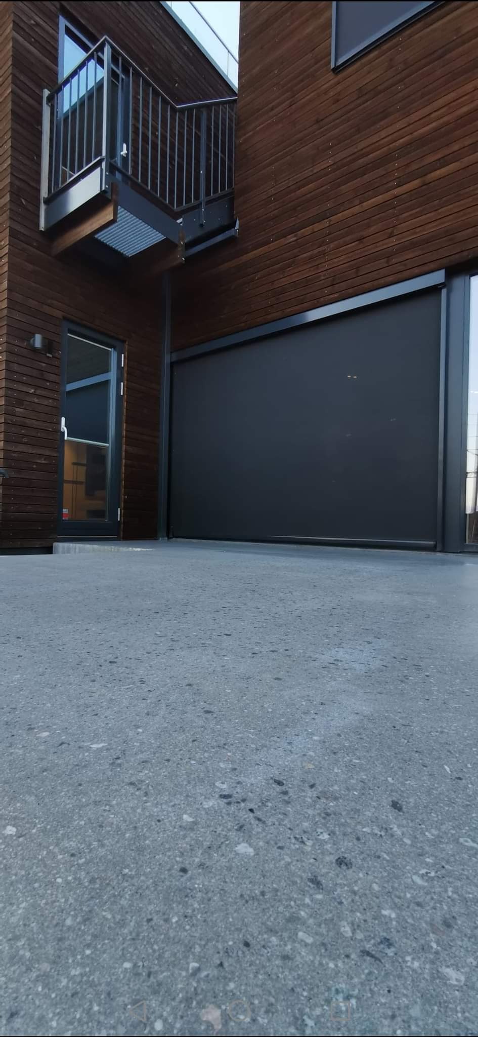 Szara, gładka powierzchnia betonowa przed budynkiem z drewnianą elewacją i balkonem z metalową balustradą.