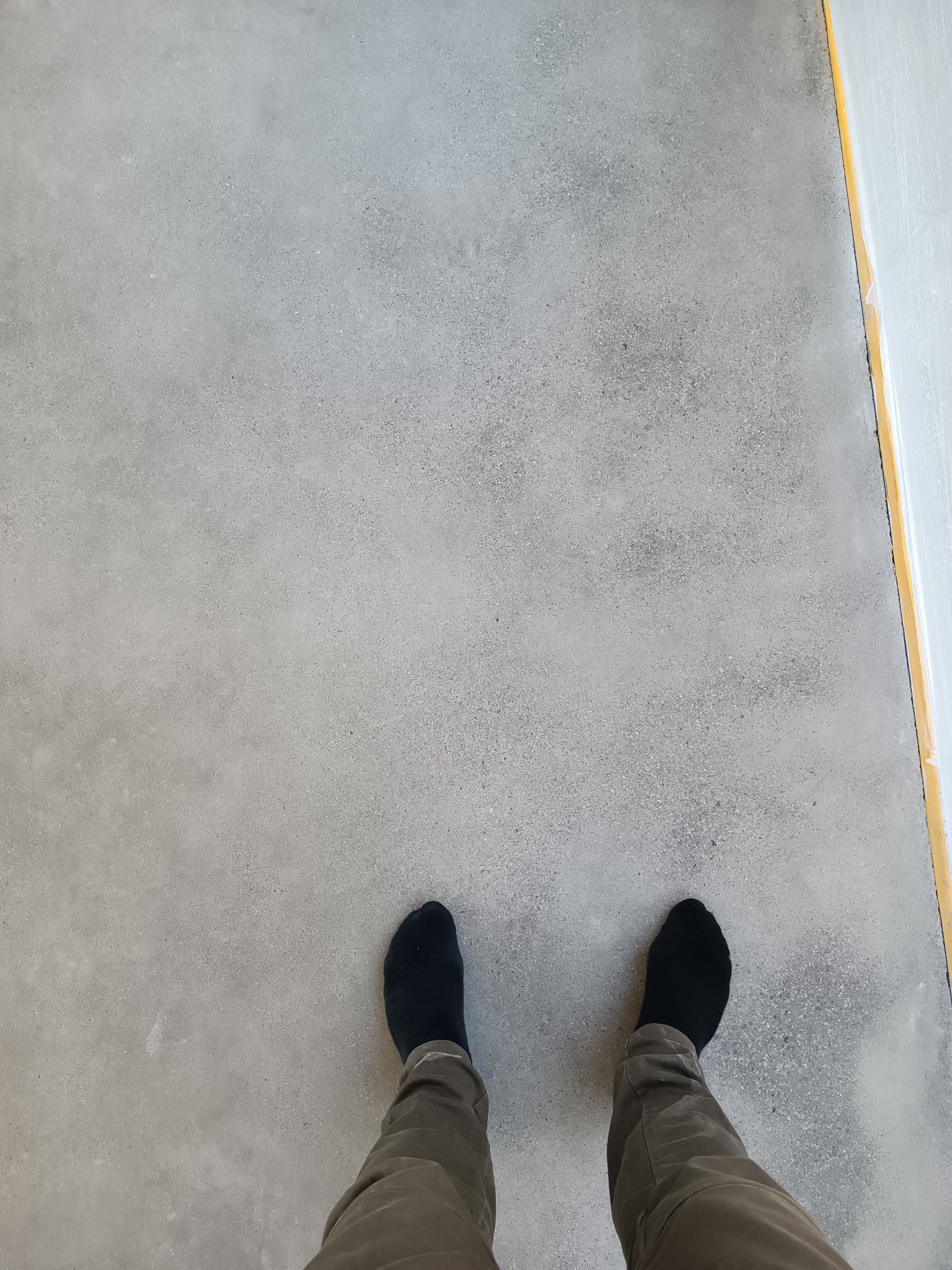 Szara, gładka powierzchnia podłogi z widocznymi stopami w czarnych skarpetach i spodniami khaki. Wzdłuż krawędzi widoczna żółta taśma malarska zabezpieczająca ścianę.