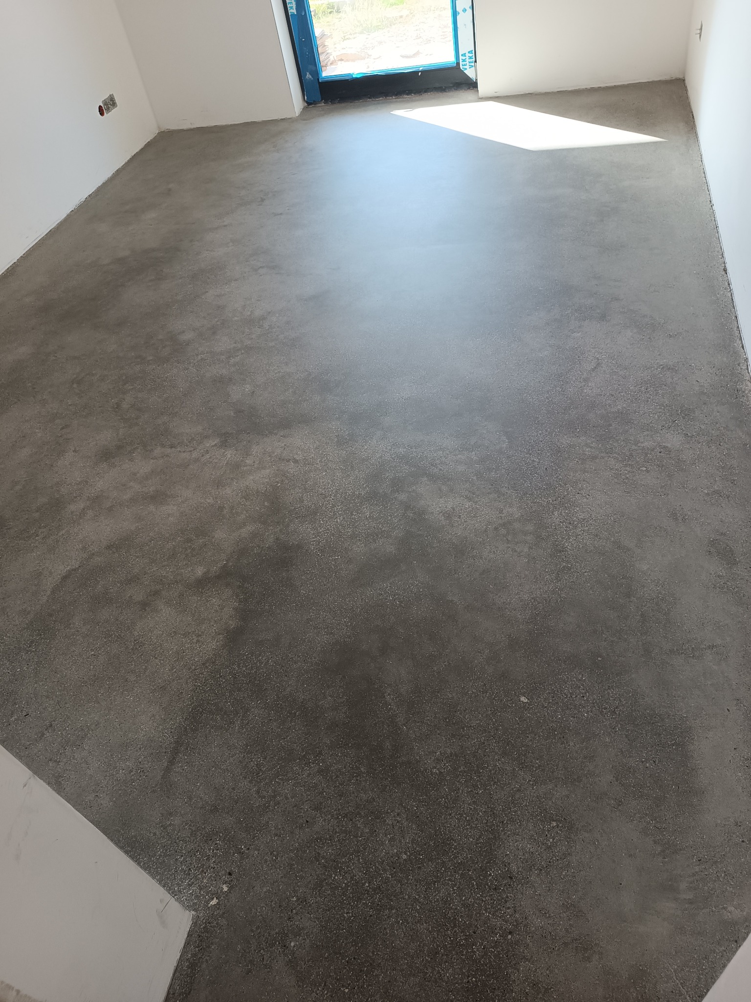 Szara, gładka powierzchnia wylewki w nowym pomieszczeniu z białymi ścianami i oknem w tle, widoczne ślady po procesie wygładzania.