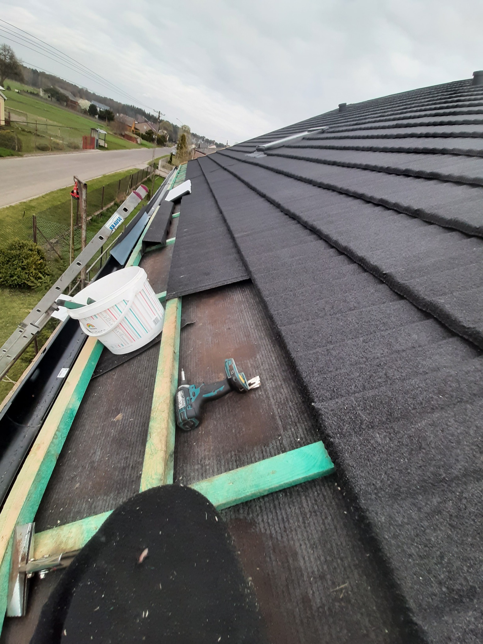 Montaż pokrycia dachowego z użyciem wkrętarki akumulatorowej, widoczne elementy konstrukcyjne dachu, rynna, drabina i wiadro z farbą w tle.