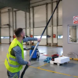 Pracownik w kamizelce odblaskowej używa długiego kija do mycia okien w hali w Gralewie, widoczny sprzęt do mycia i znak zakazu palenia.