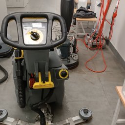 Clean Case Sp. z o.o. - Szorowarka do podłóg Karcher w trakcie pracy w biurze, widoczne ślady wody na szarej podłodze, w tle inne maszyny czyszczące i meble biurowe.