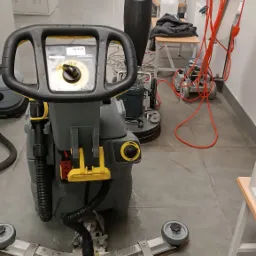 Szorowarka do podłóg Karcher w trakcie pracy w biurze, widoczne ślady wody na szarej podłodze, w tle inne maszyny czyszczące i meble biurowe.